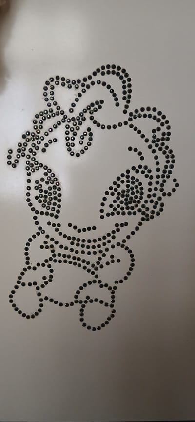 The image displays a pattern of small, dark, faceted embellishments arranged on a white, flat surface, likely fabric or paper. The embellishments are clustered together to form a design that resembles a stylized face or mask with prominent eye-like shapes and a curved line suggesting a mouth or chin. The pattern is intricate and detailed, with individual embellishments closely spaced to create the overall shape. The lighting casts subtle shadows, highlighting the three-dimensional quality of the embellishments. The image is cropped and does not show any environmental context. There is no information about the location in Andijan, Uzbekistan, as it is not depicted in the image.