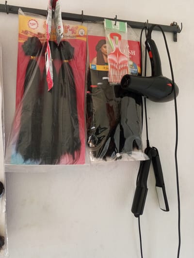 In Kabong, Nigeria, an indoor scene displays hair products and styling tools hanging on a wall. To the left, a package of black hair extensions is visible, with three bundles tied together and a pink card backing. Next to it, another package of hair extensions, also black, is partly obscured by a plastic bag containing red and green styling combs. To the right, a black hairdryer with red buttons hangs, connected to a power cord. Below the hairdryer, a black flat iron is also suspended by its cord. The wall behind the items is a plain, off-white surface, showing minor staining and texture. The overall setting suggests a retail or personal grooming space.
