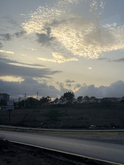 The image depicts a scene in Pampatar, Venezuela, characterized by a cloudy sky at sunset. In the foreground, a road with a yellow curb is visible. Beyond the road, a dark, overgrown hillside is dotted with debris. On the hillside, a white sign with black text reads "PROPIEDAD PRIVADA" and a phone number "0414-357.69.72." Further back, a row of houses with red tile roofs and white walls stretches across the landscape, interspersed with palm trees and other vegetation. A cell tower stands tall among the trees. In the distance, a multi-story building is visible, suggesting a more urbanized area. The overall impression is one of a semi-urban environment during twilight.