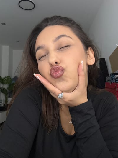 A close-up portrait shows a young woman posing indoors. The image is initially rotated 90 degrees counter-clockwise. The woman, appearing to be in her 20s or early 30s, has long, dark hair. She is wearing a black long-sleeved top.
Her eyes are closed, and her lips are pursed in a "duck face" or puckered pose, conveying a playful or expressive mood. Her right hand is raised, with fingers slightly spread, touching her chin and lower jawline. On her right ring finger, she wears a silver-toned ring featuring a floral or snowflake-like design adorned with multiple light-colored stones. Her fingernails have a neat, light-colored manicure. A delicate necklace with a small, sparkly cross pendant is visible on her chest.
The background features a plain grey wall. In the upper background, a dark object with white text is partially visible, which includes "CRA" and "ENERGY DRINK." Towards the lower right, a green leafy plant is partially visible. Another dark, circular object is on the wall in the lower left. The lighting is even and bright, consistent with an indoor setting. The scene captures a casual, posed moment, likely a selfie. While the location context is Mönchengladbach, Germany, no specific visual cues within the image confirm this.