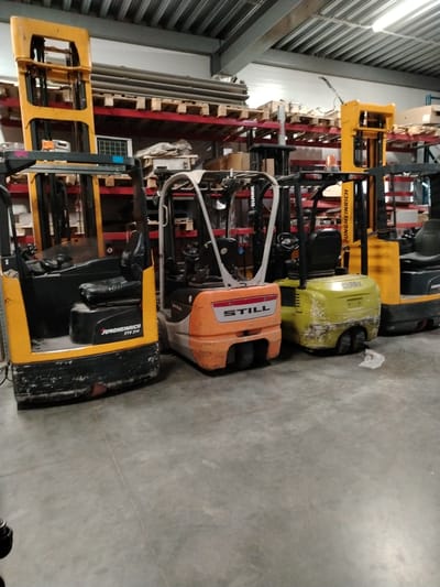 The image displays an indoor warehouse or industrial storage facility, primarily featuring a row of parked forklifts. Four forklifts of varying models and brands are visible on a concrete floor in the midground.
From left to right, the first forklift is a yellow and black Jungheinrich reach truck, with "JUNGHEINRICH ETV 214" visible on its side. It has a tall mast extended upwards. Next to it is a smaller, orange and black forklift, branded "STILL". Following this is a compact, green-yellow and black forklift with "CLARK" written on its rear. The last visible forklift on the right is another yellow and black Jungheinrich model, positioned further back. All the vehicles exhibit signs of heavy use, with scuffs, dirt, and wear on their bodies.
In the background, tall, red metal shelving units stretch across the frame, filled with numerous wooden pallets. Some pallets are empty, while others hold cardboard boxes and various packaged items. The shelving extends high up towards the ceiling, which is composed of corrugated metal and features visible structural beams and overhead lighting fixtures. The concrete floor shows some dirt and debris, consistent with an industrial environment. No people are present in the scene, and no specific activity is taking place; the forklifts appear to be parked. The setting is brightly illuminated by internal lighting, suggesting an operational warehouse at any time of day.