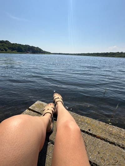 This media file features a close-up shot of a person's legs and feet, adorned with light-colored sandals, resting on a wooden dock. The dock appears to be covered with wire mesh in some areas. The background is dominated by a tranquil body of water, reflecting the bright sunlight, which creates a shimmering effect. Further in the distance, a lush green shoreline with dense trees borders the water, suggesting a natural, perhaps wooded, environment. The sky above is a clear, vibrant blue, indicating fair weather. The sunlight suggests it's daytime, likely mid-morning or afternoon. The overall mood is relaxed and peaceful, evoking a sense of leisure and enjoyment of the outdoors. There is no visible text in the image. The location context provided, Great Yarmouth, United Kingdom, hints at a coastal or lakeside setting within that area.