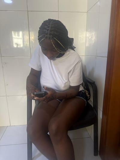 In an indoor location in Ikate, Nigeria, a female is depicted seated on a dark office chair with a black cushion. She has dark skin, black braided hair with prominent blonde strands, and wears a white short-sleeved t-shirt and dark patterned shorts. Dark, intricate henna-like patterns are visible on both forearms. She holds a dark-colored smartphone horizontally with both hands, directing her gaze downwards towards the device. The background consists of white ceramic tiles on the wall, showing a reflection of a window structure on the upper left. A light brown wooden door or door frame is partially visible on the right side of the frame.