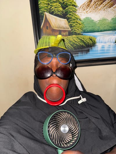 The image shows a close-up, top-down view of a person, likely relaxing indoors. The individual has dark skin and is wearing a black hoodie or similar garment. A bright yellow mesh cap is visible on their head. They are sporting two pairs of glasses: one with distinct blue frames and another pair with dark lenses, placed over the blue-framed ones. A prominent red, ring-like object, possibly an ear cuff or a large earring, is worn on their right ear.
A small, portable fan with a green exterior and black blades, which appear to be dusty, is positioned near the person's head, suggesting it is in use. A white plastic clip or tag is attached to the black clothing. The background features a cream-colored wall and a large framed painting. The artwork depicts a scenic landscape with a large waterfall, lush green trees and foliage, and what appears to be a light-colored wooden structure or hut.
The overall scene suggests an indoor setting, possibly in Yelwa, Nigeria, during warm weather, indicated by the use of the fan. The lighting appears to be ambient indoor light. No discernible text is visible in the image, and the person's expression, from the partial view, appears calm.