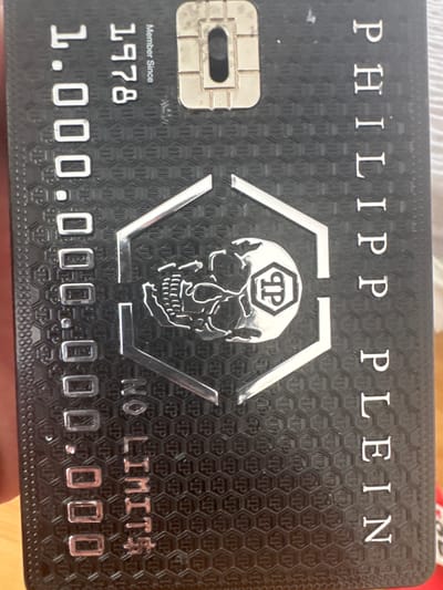The image shows a close-up, vertical view of a black membership card with metallic silver lettering and details. The card features the number "1.00.000.000" in a large font on the left side, followed by "NO LIMIT$" below it. The top right corner has a chip. The name "PHILIP PLEIN" is written vertically down the right side of the card. In the center, there is a hexagonal outline enclosing a skull graphic with a shield-like symbol superimposed on its forehead. The card has a textured, honeycomb-like pattern in the background. The card is held in a person's hand, partially visible at the bottom left. There are no visible indications of the city of Schkopau, Germany, or any environmental context.