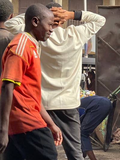 Two men are sitting in close proximity. One wears an orange shirt with white stripes on the shoulders and black shorts. The second man behind him wears a white long-sleeved shirt with a black wrist cuff and dark gray pants. The back of his head and hand are visible over the first man's head. The men appear to be in a transport vehicle or cart with various items in the background. The image is rotated slightly.