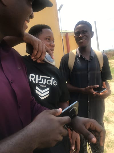 The scene depicts three young adult males engaged in a casual outdoor interaction in Uyo, Nigeria, under bright daylight.
The individual in the left foreground, partially visible, wears a dark purple long-sleeved shirt and a black baseball cap. Their face is mostly out of frame, revealing a smiling mouth. A right hand extends from this individual, resting on the shoulder of the central figure and holding a dark smartphone featuring a prominent circular camera module. Another hand, likely belonging to the same person, is visible in the lower left, holding a similar dark smartphone.
The central individual wears a black t-shirt with "RHHude ClassiGLIFE" and a white chevron design printed on the front. This person has short dark hair and is oriented slightly to the right, looking away from the camera. A white cylindrical object is held in their left hand near the waist.
The third individual stands in the right background, wearing a dark grey/black short-sleeved collared shirt with a subtle grid pattern and a light tan backpack. This person has short dark hair and is smiling broadly, directly at the camera. Their hands are positioned in front, holding a dark smartphone with a silver edge.
The background includes a light yellow building wall on the left, with a segment of a green roof visible above it. A solar-powered street light pole extends upwards on the left, and a wooden utility pole is present on the right. The ground is composed of light-colored dirt with scattered patches of green grass. The sky overhead is bright and clear, consistent with an outdoor daytime setting. All individuals are holding or interacting with smartphones.