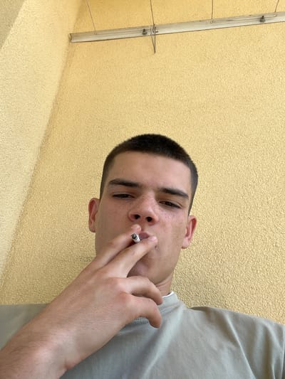 A young person with short dark hair and freckles is pictured close-up, holding a cigarette to their lips. They are wearing a light-colored t-shirt. The background is a textured yellow wall, with a metal fixture and wire visible on the left side, suggesting an outdoor setting. The lighting suggests it is daytime. The person's gaze is directed towards the camera, and their expression is neutral. The image is oriented vertically, with the person's head at the top and their torso extending downwards. The overall impression is casual and informal. There is no visible text in the image.