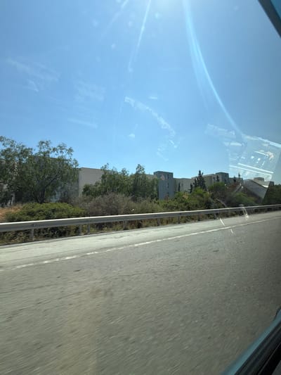 The image captures a daytime view of a road and buildings in Parekklisia Community, Cyprus. The foreground features a section of asphalt road with white lane markings, moving from bottom right to top left. A silver metal guardrail runs parallel to the road, separating it from a vegetated area. Behind the guardrail, there are various types of trees and bushes, some appearing dry and others lush green.
Further in the background, a complex of multi-story buildings is visible. These structures are modern in design, with light-colored facades and visible windows. The buildings are interspersed with more greenery, including tall, slender trees. The sky above is clear and blue, with prominent lens flare and reflections on the surface from which the photo was taken, likely a vehicle window. The overall impression is one of a suburban or semi-urban area with mixed residential and natural elements. There are no people or animals visible in the scene.