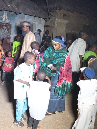 A group of people, mostly children, surround an adult woman outdoors. The woman wears a head wrap and a green, red, and black shawl. She holds a phone in her hand. The children wear colorful clothing, including shirts and pants in shades of blue, green, red, and white. Some individuals stand near buildings with weathered walls and corrugated metal roofs. The lighting suggests it is evening or nighttime.
