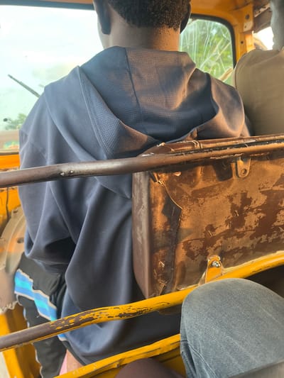 The image captures an interior view of a yellow vehicle, possibly a tuk-tuk, in Kuchingoro, Nigeria. The primary subject is a person wearing a dark blue shirt, their head and shoulder visible in the foreground. The person appears to be sitting or leaning within the vehicle.
The interior of the vehicle shows signs of wear and tear. A rusty, metallic object, possibly a part of the vehicle's structure or a cargo holder, is prominently visible, its surface exhibiting a mottled brown and beige texture with signs of chipping and staining. Yellow painted metal bars and tubes are also part of the vehicle's interior, some with peeling paint. A section of dark grey fabric, likely upholstery or clothing, is visible on the right side of the frame. A broken or cracked transparent material, possibly a window or windshield, is seen through the opening on the left, revealing a blurred green, outdoor scene consistent with daytime.
The overall impression is one of a functional, perhaps aging, mode of transport in a busy environment. The lighting suggests it is daytime. There are no discernible signs of text in the image. The scene is likely a snapshot of daily life, possibly a journey or a pause during transport.