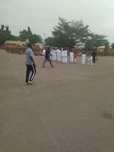 The scene takes place on a paved, grey, outdoor area in New Karu, Nigeria, located at coordinates 8.993, 7.571. A group of people, predominantly young children, are in the middle of the scene, all wearing white martial arts uniforms. They are arranged in lines, facing towards the right of the image, and appear to be participating in a martial arts training session. There are two adult figures in the scene: one standing on the left, looking directly at the camera, and one standing to the right of the children, facing the group. The children appear to be following the instructions of the adult standing to the right, who is likely their instructor. The adults are both wearing dark clothing, while the children are all wearing white uniforms. The environment is open and paved, with a green field on the left and buildings in the background. The weather appears overcast, with a muted sky. The paved area appears relatively empty, with only a few small objects scattered on the ground. The image is taken from an elevated viewpoint, and the focus is on the group of people in the middle of the scene.