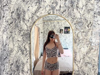 A young woman is taking a mirror selfie in a bedroom. She is wearing a leopard print pajama set consisting of a bralette and shorts. The woman has long brown hair and is holding a smartphone to take the picture. She has a tattoo on her left arm. In the reflection of the mirror, a bed with pink pillows and a patterned wallpaper can be seen. The room appears to be located in an apartment or house in Blizhnii Khutor, Moldova. The overall lighting is bright, suggesting it is daytime.