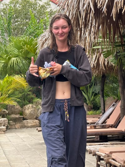 A young person with wet, shoulder-length brown hair is standing outdoors, giving a thumbs-up and holding a small object wrapped in gold foil in their left hand. They are wearing a dark gray zip-up hoodie, a black crop top, and dark blue baggy pants with a braided belt featuring red, yellow, and brown tassels. The person is smiling, looking towards the camera. The background includes lush green foliage, large rocks, and thatched roofing. Several lounge chairs are visible in the middle ground. The ground is paved with light-colored square tiles. The location appears to be a resort or a park-like setting within Nam Hoa Lư Ward, Vietnam.