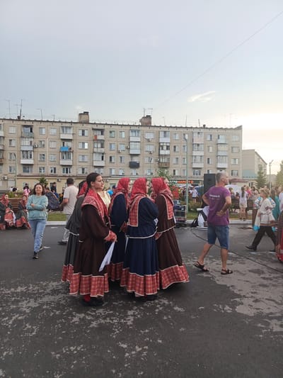 This media file captures an outdoor gathering in a residential area of Krivodanovka, Russia. The scene is set against a backdrop of a typical Soviet-era apartment building, characterized by its monotonous facade and numerous balconies. The sky appears overcast, suggesting it might be late afternoon or early evening, and the weather seems mild.
The primary subjects are a group of people, many of whom are dressed in traditional Russian folk costumes. These costumes feature long, full skirts and ornate embroidery, with women wearing headscarves. The people are standing and walking on an asphalt surface, possibly a street or a public square.
The activity appears to be a cultural event or festival. Some individuals are posing for photographs, while others are engaged in conversation. In the background, there are hints of a stage or performance area with sound equipment, and other attendees are visible, some seated on benches. One notable detail is a sign with Cyrillic text, although it is not entirely legible. A man in a purple t-shirt with Cyrillic text and a woman in jeans and a teal sweatshirt are also prominent figures in the foreground, observing the scene. The overall atmosphere seems relaxed and festive, with a sense of community engagement in a cultural celebration.