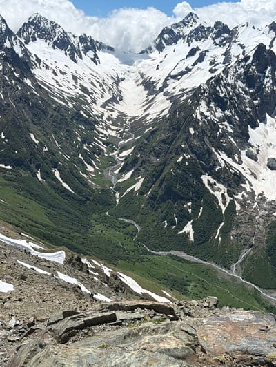 The image depicts a vast, rugged mountain landscape in the vicinity of Верхне-Тебердинское сельское поселение, Russia. Dominating the scene are towering peaks, partially covered in snow and ice, with dark, rocky outcrops interspersed with patches of white. A deep valley carves through the mountains, its slopes densely covered in lush green vegetation, suggesting a subalpine or alpine environment. A winding river or stream is visible flowing through the valley floor, its path marked by a lighter, reflective surface. The foreground is characterized by loose scree and large rocks, indicating a high-altitude, rocky terrain. The sky above is a mix of bright blue with scattered, fluffy white clouds, suggesting fair weather. The overall impression is one of immense natural beauty and wilderness, typical of a remote mountainous region. There are no people or man-made structures clearly visible in the image.