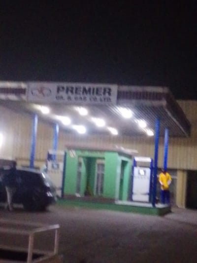 The image depicts a nighttime scene at a Premier Oil & Gas Co. Ltd. service station in Babban Giji, Nigeria. A brightly illuminated canopy, supported by blue pillars, covers the foreground area. Underneath the canopy, a dark-colored vehicle is positioned on the left side. To the right, a white fuel pump dispenser is visible. Behind the pump, a small, green building with several door-like openings is situated. One individual, wearing a yellow shirt and blue trousers, stands near the fuel pump on the right. The ground is dark asphalt, and a light-colored, rectangular object is partially visible in the lower-left foreground. The background is dark, indicating night, with faint outlines of what appear to be structures.