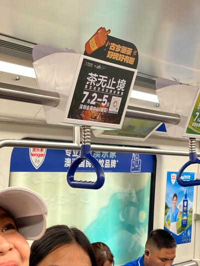This image appears to be taken inside a subway car in Futian District, China. The primary subjects are three individuals, two of whom are partially visible in the foreground. One person is wearing a blue shirt and has dark hair, with a black earbud in their ear. Another person is wearing a light-colored baseball cap. The background features advertisements and handrails typical of public transportation.
One prominent advertisement is for a "Tea Fair" showcasing "Ancient and Modern Trendy Tea" that is "Fun and Interesting." Another advertisement, with the text "Tea Without Borders," promotes a "Tea Culture Art Immersive Exhibition" taking place from July 2nd to July 5th at the Shenzhen Convention and Exhibition Center (Futian). The word "TEA" is also visible on a graphic of a teacup.
Another advertisement, featuring the brand "澳尔禾 Aerogard," is also visible. The overall scene suggests a commercial or event promotion within a public transport setting. The lighting indicates it could be daytime, but there are no clear weather cues. The presence of the advertisements and the overall environment suggest a vibrant urban setting.
