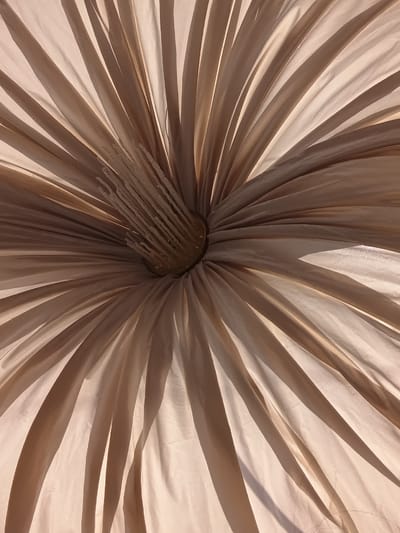 An underside view of a light-colored, fabric umbrella reveals its internal structure. The fabric is pleated and fanned out from a central point, creating a radial pattern of light and shadow. At the center, a metallic mechanism with string-like supports is visible. The lighting suggests an outdoor setting, possibly under sunlight, casting distinct shadows on the fabric. The image does not depict any urban or environmental features specific to the city of Osh, Kyrgyzstan.