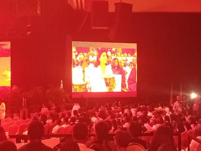 The image shows a large indoor gathering in Itirin, Nigeria, likely an event or conference, with many seated attendees facing a large screen displaying a video. The screen shows a woman in a white outfit and other people in the background, possibly related to music or cultural performance, as indicated by the presence of traditional drums. The room is bathed in red lighting, possibly from stage lights, and there are speakers visible above the screen. A few individuals are standing on the periphery, including one with a camera or recording device. The seating area is filled with people, largely facing the screen.