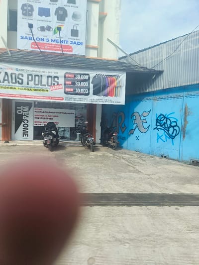 The image shows a storefront in Purwakarta, Indonesia, located at the coordinates -6.538 latitude and 107.441 longitude. The storefront is selling polo shirts and has a sign advertising "Kaos Polos" (polo shirts) with a price list. The storefront is open and there are two motorcycles parked in front. The building next door is a light blue color and has graffiti on it. The street is paved and appears to be in a residential area. The image is partially obscured by a finger in the foreground.