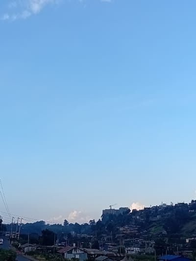 An upward-angled outdoor shot exhibits a vast, clear blue sky occupying the left side of the frame. The right side features a hillside densely populated with houses and buildings. A construction crane is visible among the structures. Power lines run along the base of the hill near a road with a single vehicle. The overall scene suggests a settlement built on a steep slope under a cloudless sky.