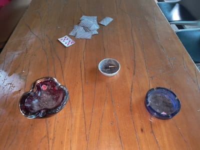 The image displays a wooden table with a scattered deck of playing cards, two ashtrays, and a small metal container with ash and a cigarette butt. One ashtray is made of dark red glass and is shaped like a heart. The other ashtray is made of blue glass and is circular. The cards are face down except for a single card showing the 10 of hearts. The metal container, possibly a small can or lid, contains ash and a single cigarette butt. The table surface shows wear and tear with visible wood grain and scratches. In the background, out of focus, are parts of chairs, suggesting an indoor setting. The lighting indicates it could be daytime. The scene appears to be in Rivne, Ukraine. There is no visible text other than a small sticker on the red ashtray which is illegible.