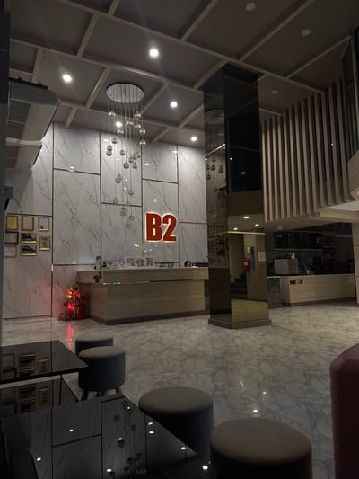 The image depicts the interior of a modern hotel lobby, identified as the B2 Surat Thani City Hotel. The scene is captured from a low angle, looking towards a reception desk and a coffee shop area.
The lobby features a sleek, contemporary design with marble-patterned flooring and walls. A prominent "B2" logo is illuminated on the back wall behind the reception desk. Suspended from the coffered ceiling is a modern chandelier composed of multiple glass orbs.
To the left of the reception desk, several framed certificates or awards are mounted on the wall. A small, decorative red Christmas tree with lights stands near the reception desk. The reception desk itself is a light-colored wooden structure. A person is partially visible behind the desk, attending to the reception.
To the right of the reception area, a glass-encased pillar reflects the surrounding environment. Further to the right is a coffee shop counter with a menu board displaying drinks and food items. The coffee shop area is branded as "WAKE UP". Seating arrangements, including round, upholstered stools and reflective coffee tables, are visible in the foreground. The overall lighting is soft and ambient, creating a welcoming atmosphere. The location is the B2 Surat Thani City Hotel, situated in Surat Thani City Municipality, Thailand.