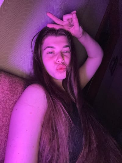 The image shows a young woman lying on her side, looking at the camera. She has long, brown hair and is making a kissy face. Her eyes are open, and she is wearing a black top. She is holding up her right hand in a peace sign. The scene appears to be indoors, with purple lighting. The background is dark, with a hint of what looks like a couch or bed. The woman's skin is illuminated by the purple light, and there are some small marks visible on her arm. The overall mood of the image is playful and casual. The location context is Мещёры, Russia.