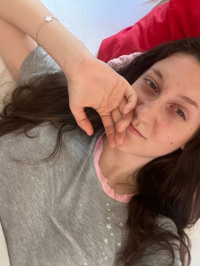 A young woman with long, dark, wavy hair is lying down, looking towards the camera. She is wearing a gray t-shirt with a pink collar. Her left arm is bent, and her hand is resting on her face. She has a star-shaped bracelet on her wrist. The background appears to be a bed with white and red bedding. The scene is indoors, likely in a bedroom. There is no information available to suggest the location is in the city of Popintsi, Bulgaria.