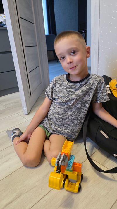 A young boy is sitting on a light-colored wooden floor, playing with a construction toy. He is wearing a gray camouflage t-shirt and green shorts, with striped socks. The toy is made of brightly colored plastic blocks, predominantly yellow and orange, with some blue and gray pieces. To his left, a black backpack is visible, with a yellow object on top of it. The boy is looking towards the camera with a slight smile. The setting appears to be indoors, likely a room in a house, with a light-colored wall in the background. The image is taken from a slightly elevated angle, looking down at the boy. There is no indication of the time of day or weather conditions. The location context is Bulgakovo, Russia.
