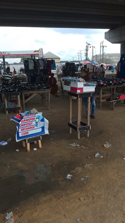 The image shows a market scene under an overpass in Effurun, Nigeria. Several wooden tables display various items for sale, including shoes and clothing. A sign advertising financial services (transfer, deposit, recharge card) sits on a small stool. Several people are visible, including vendors and potential customers. In the background, there is a gas station with "BAY OIL" visible. The ground is dirt, with scattered litter and debris. The sky is overcast. Power lines and poles are also visible in the background.