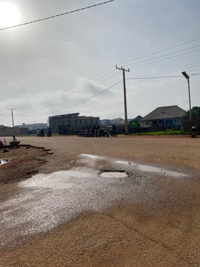 The image depicts a street scene in Kwang, Nigeria, likely in the late afternoon given the overcast sky and diffused sunlight. The perspective is from ground level, looking towards a row of buildings and a street.

The primary subjects are the road, buildings, and several motorcycles. The road surface is asphalt, characterized by a rough texture and the presence of several puddles, suggesting recent rain or damp conditions. One large puddle with a distinct rectangular shape is particularly prominent, reflecting the sky. There are also several potholes and rough patches on the road.

In the background, a line of buildings of varying heights and architectural styles can be seen. Some appear to be residential or commercial structures, with multiple stories and balconies. One building is under construction. Amidst the buildings, green vegetation is visible, indicating a potentially tropical or sub-tropical environment.

Several motorcycles are parked or being ridden along the road, forming a small procession. People are visible on and around the motorcycles, though their features are not clearly discernible due to the distance and resolution. They appear to be dressed in everyday clothing.

Overhead, power lines crisscross the sky, supported by utility poles. A lamppost is also visible. The sky is cloudy and a bright, hazy sun is partially visible in the lower left corner. The overall impression is one of an active, everyday street scene in a Nigerian town. There is no visible text in the image.