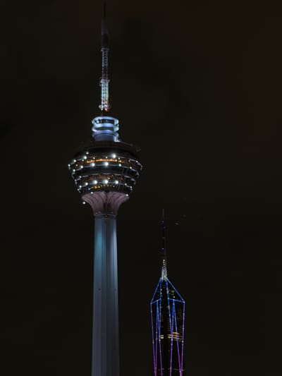 The image shows two illuminated towers at night, likely in Kuala Lumpur, Malaysia, given the location context. The foreground features a close-up of the lower section of a tower, which appears to be the Kuala Lumpur Tower (Menara Kuala Lumpur). This section is adorned with numerous bright lights, highlighting its intricate, ribbed structure. The lights are predominantly white and orange, casting a warm glow. The tower's shaft extends to the left, appearing as a smooth, metallic gray surface.
In the background, a second, more distant tower is visible. This tower has a geometric, lattice-like structure decorated with blue and purple lights, creating a more modern and angular appearance. It is significantly thinner and extends further into the dark sky. The dark background suggests it is nighttime. There are no people visible. The overall scene conveys a sense of urban grandeur and architectural beauty illuminated against the night sky. There is no visible text in the image.
