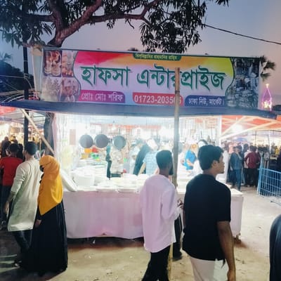 This image captures a bustling outdoor market or fair scene during dusk or evening, indicated by the artificial lighting and dim sky. The central focus is a market stall, likely selling household goods, particularly stacks of white ceramic or plastic dinnerware like plates, bowls, and serving dishes. Several dark-colored frying pans or woks are hanging prominently at the front of the stall.

Numerous people, predominantly men, are seen walking around, browsing, and interacting within the market. On the left, a woman wearing a yellow hijab and a black abaya is walking past the stall. Other individuals include a man in a white long-sleeved shirt and dark pants next to her, and a man in a black t-shirt and light-colored pants facing away from the camera on the right. The background is filled with more market-goers and other stalls, contributing to a lively atmosphere. A blue metal fence is partially visible on the right.

Above the stall, a large banner acts as signage. The text on the banner reads: "বিস্মিল্লাহির রাহমানির রাহিম" (Bismillahir Rahmanir Rahim - In the name of Allah, the Most Gracious, the Most Merciful). The main business name is "হাফসা এন্টারপ্রাইজ" (Hafsa Enterprise). Below this, it lists "প্রোঃ মোঃ শরিফ" (Proprietor: Md. Sharif) and a phone number: "01723-203506". The location is stated as "ঢাকা নিউ, মার্কেট।" (Dhaka New, Market.). Partially visible text on the left side of the banner reads "প্রয়োজনেঃ চরফ", suggesting a connection to Charfasson, Bangladesh, where the event might be taking place. Another partial text "মাসব্যাপ" (meaning "month-long" or "monthly") is visible, often associated with fairs. In the far background on the right, a distinctive light-up structure, possibly a decorative tower or spire with neon lighting, adds to the festive ambiance. A large tree with dark foliage is visible at the top of the image, overhead.