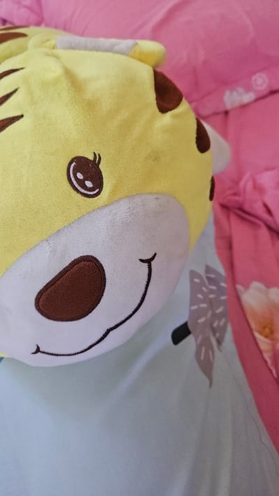 A stuffed animal with a yellow and white face lies on bedding. The left side of the face is yellow with brown spots, an embroidered eye, and brown stripes. The right side is white with a brown embroidered nose and a stitched-on smile. The bedding has a pink sheet on the left side and a blue sheet with a tree design on the right. The location is unknown.