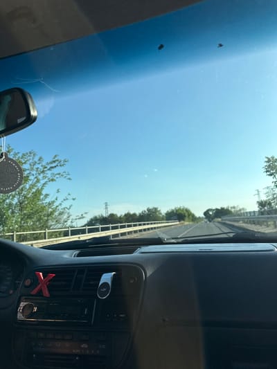 The image is taken from inside a car, looking out through the windshield. The view shows a highway with trees and utility poles lining the sides. The sky is clear and blue, suggesting a sunny day. There are some small marks on the windshield, possibly from dust or insects. Inside the car, the dashboard is visible, including an air vent with a red "X" decoration and a phone mount. The car appears to be in motion, based on the perspective of the road. The location is specified as Konstantinovo, Bulgaria.
