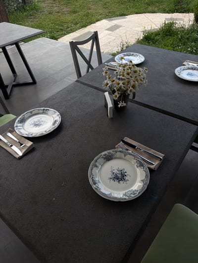An outdoor seating area is set up with a dark gray stone table and chairs. On the table are several place settings, each with a decorative plate featuring a blue floral pattern, cutlery, and a napkin. A vase filled with a bouquet of daisies sits in the center of the table. The table is situated on a tiled patio, with grass and vegetation visible in the background. The scene appears to be in Dsegh, Armenia, given the context provided.