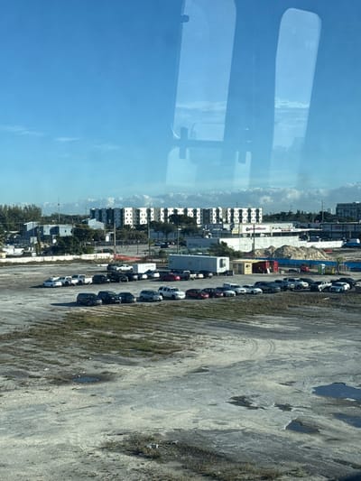 The image depicts an urban landscape in Miami, United States, captured from an elevated viewpoint through a window exhibiting reflections of two large, vertical rectangular objects. The foreground is dominated by a vast, unpaved dirt lot exhibiting patches of sparse, dry vegetation, tire tracks, and several small standing puddles.
In the midground, approximately thirty vehicles, including sedans, SUVs, and pickup trucks of various colors (predominantly black, white, red, and silver), are parked in a loose, arc-like formation on the dirt surface. Behind the vehicles are two large utility trailers: one white enclosed semi-trailer and a longer, light blue/gray enclosed semi-trailer. Adjacent to these are a small, yellow portable office structure and a red shipping container. Further back, large mounds of excavated earth or construction materials are visible.
The background features a dense collection of urban structures under a clear blue sky with minimal cloud cover. These include several low-rise commercial and industrial buildings, one of which prominently displays a "Cost Plus" sign. Behind these, a series of multi-story residential apartment or condominium buildings, characterized by white facades and dark windows, form a consistent horizontal line. Scattered trees, utility poles, and additional distant buildings complete the horizon. No individuals are visible, and no overt actions or interactions are discernible within the scene. The environment suggests an area undergoing development or mixed-use zoning within Miami.