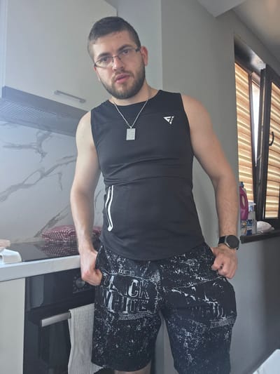 A man is standing in what appears to be a kitchen. He is wearing glasses, a black sleeveless shirt, and black shorts with white lettering. He has a beard and a necklace with a pendant. He is gripping the sides of his shorts with both hands. The kitchen has white cabinets, a microwave, a towel, and a window with blinds. The exact location in the city of Yagodovo, Bulgaria cannot be determined from the image.