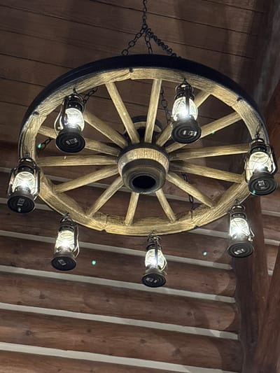 A wagon wheel chandelier with seven lantern-style lights hangs from a dark chain against a wood-paneled ceiling. The chandelier has a dark metal rim and wooden spokes. The lights are lit, casting a warm glow. The background consists of horizontal wooden planks, suggesting an interior setting. There are no people or animals visible in the image.