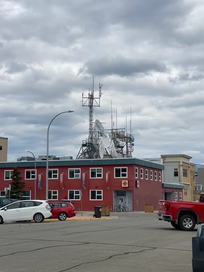 The image depicts a street scene in Whitehorse, Canada, characterized by a red building adorned with a Canadian Broadcasting Corporation (CBC) logo and a complex array of communication antennas and satellite dishes on its roof. The building is situated in an urban setting, with other buildings visible in the background, including a tan and grey structure to the right. In the foreground, a grey asphalt parking lot features several parked vehicles: a white hybrid car, a red station wagon, and a red pickup truck. Two bicycles are parked near the entrance of the red building. The sky is overcast with dark, heavy clouds, suggesting an impending or recent rainfall. The scene is captured from a street-level perspective, looking towards the building and its rooftop equipment.