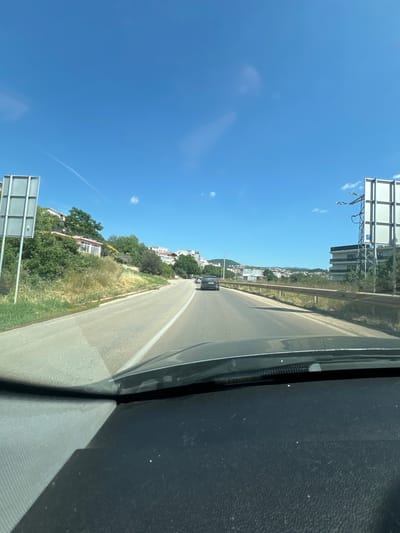 This is a first-person perspective, vertical video taken from inside a vehicle. The viewer is looking out of the passenger side window onto a road that curves away into the distance. The sky is a vibrant, clear blue with a few wispy white clouds. On the right side of the frame, the dark interior of the vehicle is visible, including part of the dashboard.

The road is asphalt with a white dividing line. There is a guardrail on the left side of the road, separating it from a grassy embankment. Beyond the embankment, there are trees and buildings, suggesting a town or village is situated on a hillside. The architecture appears to be white buildings with red-tiled roofs, consistent with Mediterranean or Balkan styles. A dark-colored car is visible driving ahead on the road.

On the left side of the road, further up the embankment, there are more buildings and lush green vegetation. A large, rectangular billboard or sign structure stands prominently near the edge of the road in the distance. The sun appears to be high in the sky, indicating it is daytime, likely late morning or early afternoon, given the bright light and clear conditions. The overall scene suggests a sunny day in a coastal or hilly region.

The location is identified as Dobra Voda, Montenegro, which is a coastal town. This context aligns with the visual cues of a sunny climate, vegetation, and buildings on a hillside overlooking what is likely the Adriatic Sea (though the sea itself is not directly visible). No people are clearly discernible in the video. The activity is simply driving through this scenic area.