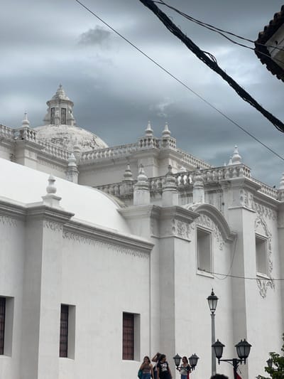 This image captures a section of the Basílica de la Natividad de María, a prominent white cathedral located in León, Nicaragua. The imposing structure features intricate white architectural details, including domes, spires, ornate carvings, and arched windows. The sky above is overcast with dark, cloudy conditions. In the foreground, several individuals are visible, appearing to be tourists or locals walking near the cathedral. One person is standing on the steps of the cathedral. Three street lamps with multiple globes are positioned in front of the building. Power lines are visible crisscrossing the sky above the cathedral.