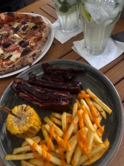 A close-up overhead shot shows a delicious meal laid out on a wooden table. The main subjects are a pizza and a plate of ribs with fries and corn. The pizza, on a white plate, is topped with ham, olives, mushrooms, and what appear to be artichoke hearts, with a slightly charred crust. Next to it, on a dark gray plate, are several racks of glazed ribs, a generous portion of golden-brown fries drizzled with an orange sauce, and a grilled corn on the cob. Two glasses filled with a clear, bubbly beverage, likely a lemonade or iced tea with mint and lime, are situated to the left of the meal, resting on white napkins. The scene appears to be an outdoor setting, possibly a restaurant patio, given the wooden table and sunlight illuminating the food. The meal seems ready to be enjoyed, suggesting a casual dining experience. There is no visible text in the image. The overall impression is one of a hearty and appealing meal, possibly enjoyed on a warm day.