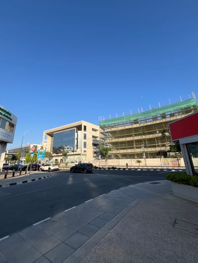 This is a landscape-oriented photograph taken outdoors in a city. The scene depicts a street with buildings under construction and adjacent modern buildings. The sky is clear and blue, suggesting a sunny day, likely late afternoon or early evening given the angle of the sun and the shadows.
In the foreground, a street runs parallel to a sidewalk with patterned paving stones. Several cars are parked or driving on the street. On the right side of the street, there's a sidewalk and then a modern building with large glass windows. Further down the street, another building is partially visible, with some greenery.
The main subject of the image is a multi-story building under construction, heavily covered in green scaffolding netting. The building appears to be made of concrete and steel. A red sign is visible at the top of this construction site. The construction appears to be a significant project, occupying a large portion of the frame.
There are no visible people in the image. Notable details include a faint crescent moon in the sky, a traffic light, and various signs along the street, some of which have text, but it's mostly illegible. One sign on the construction site appears to have safety information. Another sign on a nearby building has text which includes "Brookyn" and "Fun Through Play". The overall impression is of an urban environment undergoing development, with a mix of old and new architecture. The location is suggested to be Limassol, Cyprus.