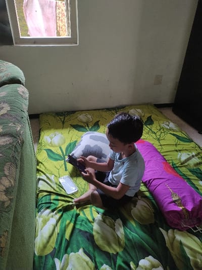 A young boy sits on a bed with a floral print bedspread. He is wearing a blue shirt and shorts. He is holding a black smartphone in his right hand and looking at the screen. Next to him is a grey pillow and a long purple pillow with a white and gold pattern. There is a white rectangular window in the white wall behind him. This scene is located in the city of Kecamatan Lawang, Indonesia at the coordinates: -7.828 latitude and 112.701 longitude.