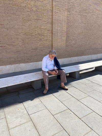 The scene depicts an elderly male seated on a stone bench outdoors, under bright daylight, in an urban setting. The location is consistent with the external walls of the Vatican City in Rome, Italy, possibly along Viale Vaticano.
The male, with grey hair and a full grey beard, wears a white long-sleeved shirt, light brown trousers, and beige slip-on loafers. A dark blue jacket is draped over his left shoulder. He is looking down at a smartphone held in both hands. He is seated on a long, light-colored stone bench with visible wear and tear, supported by darker, rough-hewn stone blocks.
Behind the bench stands a tall, weathered brick wall, predominantly light brown and yellowish in tone, constructed with horizontal stretcher bond courses. A vertical architectural element resembling a pilaster or recessed column is visible in the brickwork to the left of the man. A plinth of lighter, rough stone runs along the base of the brick wall, directly beneath the bench.
The ground is paved with rectangular grey stone slabs, characteristic of city sidewalks or plazas. Strong sunlight casts clear shadows on the ground and bench. Small pieces of refuse, including what appears to be a discarded cigarette butt, are visible on the paving stones near the bench. No other individuals are present, and there are no discernible interactions.