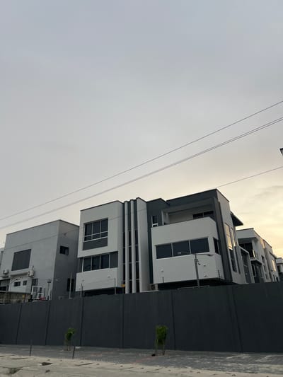 The image captures a modern urban or suburban street scene in Alaguntan, Nigeria, viewed from a low angle looking towards multi-story buildings and the sky. The sky, which dominates the left side of the frame, is hazy and light gray, subtly transitioning to a brighter, yellowish-white tone towards the upper right, suggesting late afternoon or early evening light. Several thin, dark utility lines stretch horizontally across the upper portion of the sky.
On the right side of the frame, a cluster of contemporary buildings displays a striking geometric design. They feature predominantly white and light gray facades, contrasted with dark gray or black accents, particularly in window frames and recessed architectural elements. The buildings incorporate large, dark-framed windows and what appear to be balconies or deep overhangs. An external air conditioning unit is visible on a lower section of one building.
In the immediate foreground on the right, a dark gray perimeter wall runs along the edge of what appears to be a paved or tiled ground surface, showing some texture and slight wear. A small, sparse green plant or tree sapling stands near this wall. In the background, behind one of the walls, the faint outline of a basketball hoop can be seen. The scene is devoid of people or any direct activity, presenting a static and modern environment. No visible text is present in the image.