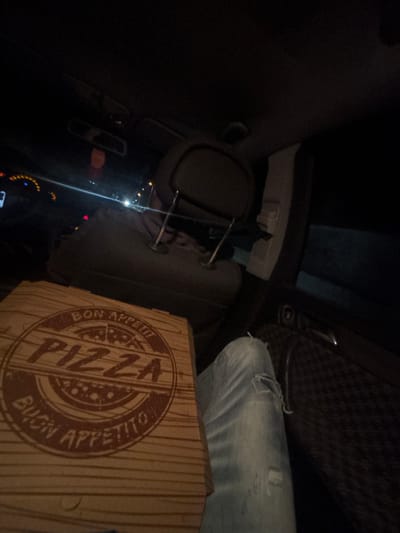 The image captures the interior of a vehicle at night, viewed from a passenger's perspective. In the immediate foreground, a rectangular cardboard pizza box with a printed wood grain texture is visible. Its lid features a circular, reddish-brown logo displaying "BON APPETIT!!!" at the top, "PIZZA" prominently in the center, and "BUON APPETITO" at the bottom. To the right of the pizza box, the upper thigh and knee of an individual's leg, clad in distressed light blue denim jeans, are partially visible.

In the mid-ground, the dark grey headrest of the front passenger seat is centrally located, supported by two chrome-colored rods. Behind the headrest, faint outlines suggest the presence of a person in the driver's seat. To the far left, a portion of the dashboard and steering wheel is discernible, illuminated by dim amber and white lights from the instrument cluster. An orange air freshener hangs from the rearview mirror, which is visible in the upper-center of the frame. The right-side passenger door panel, featuring dark, patterned upholstery, is partially visible. External light sources, appearing as blurred streaks and bright points, are visible through the vehicle's windows, indicating an outdoor roadway environment at night. The scene is located in Касапите, Bulgaria.